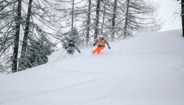 Two skiers in deep snow in front of trees.